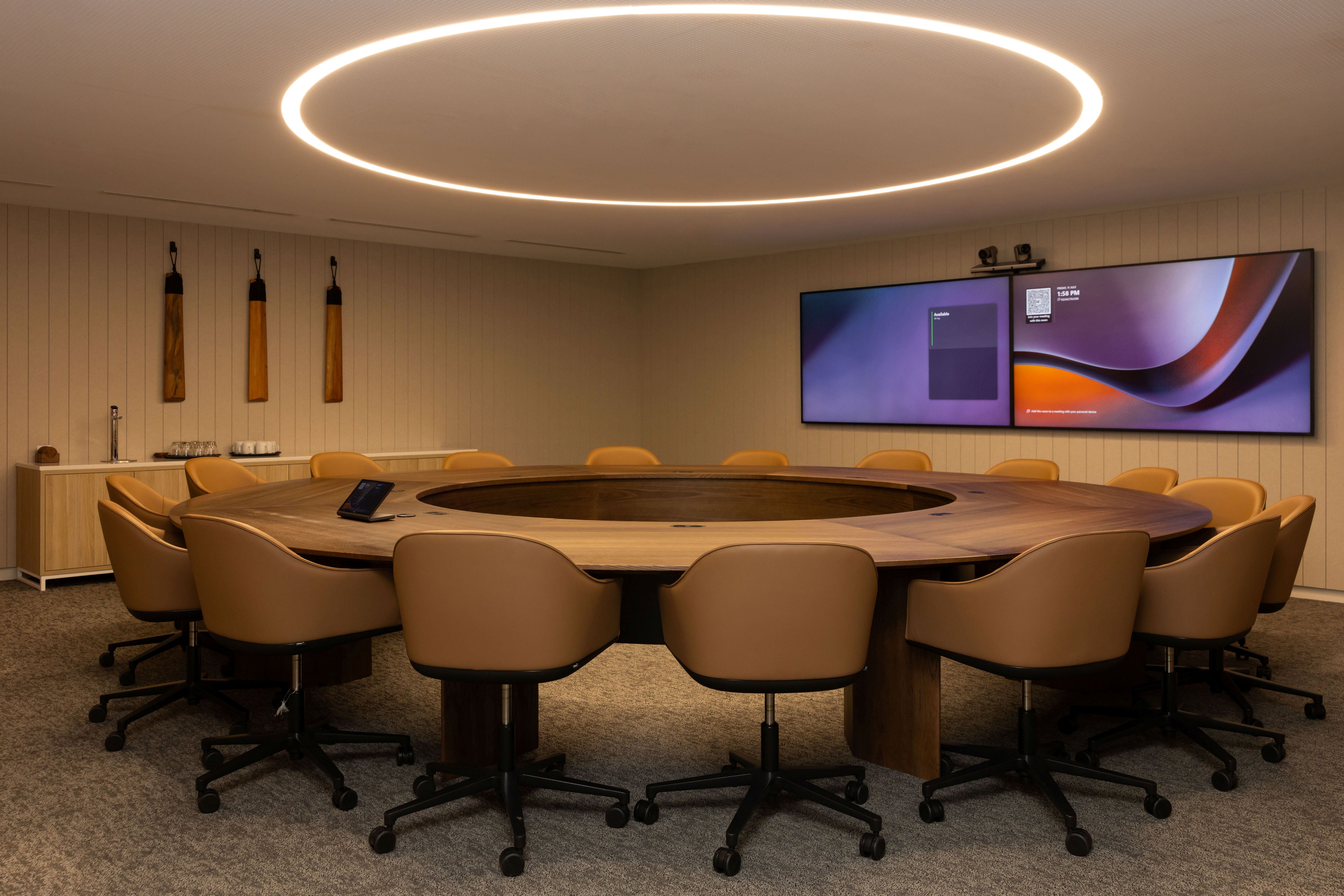 Modern conference room with round wooden table and beige chairs, featuring a large screen on the wall.