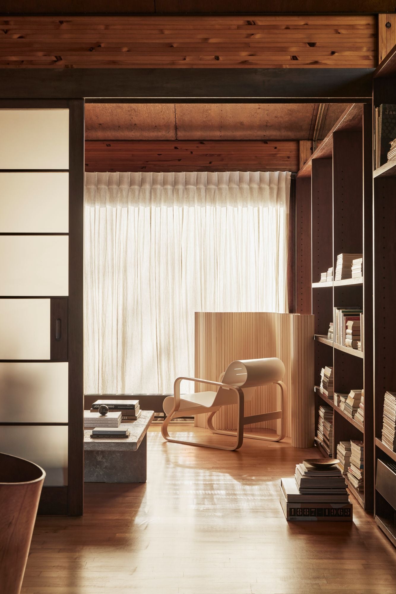 Modern interior with wooden ceiling, bookshelf, and chair in a well-lit room.