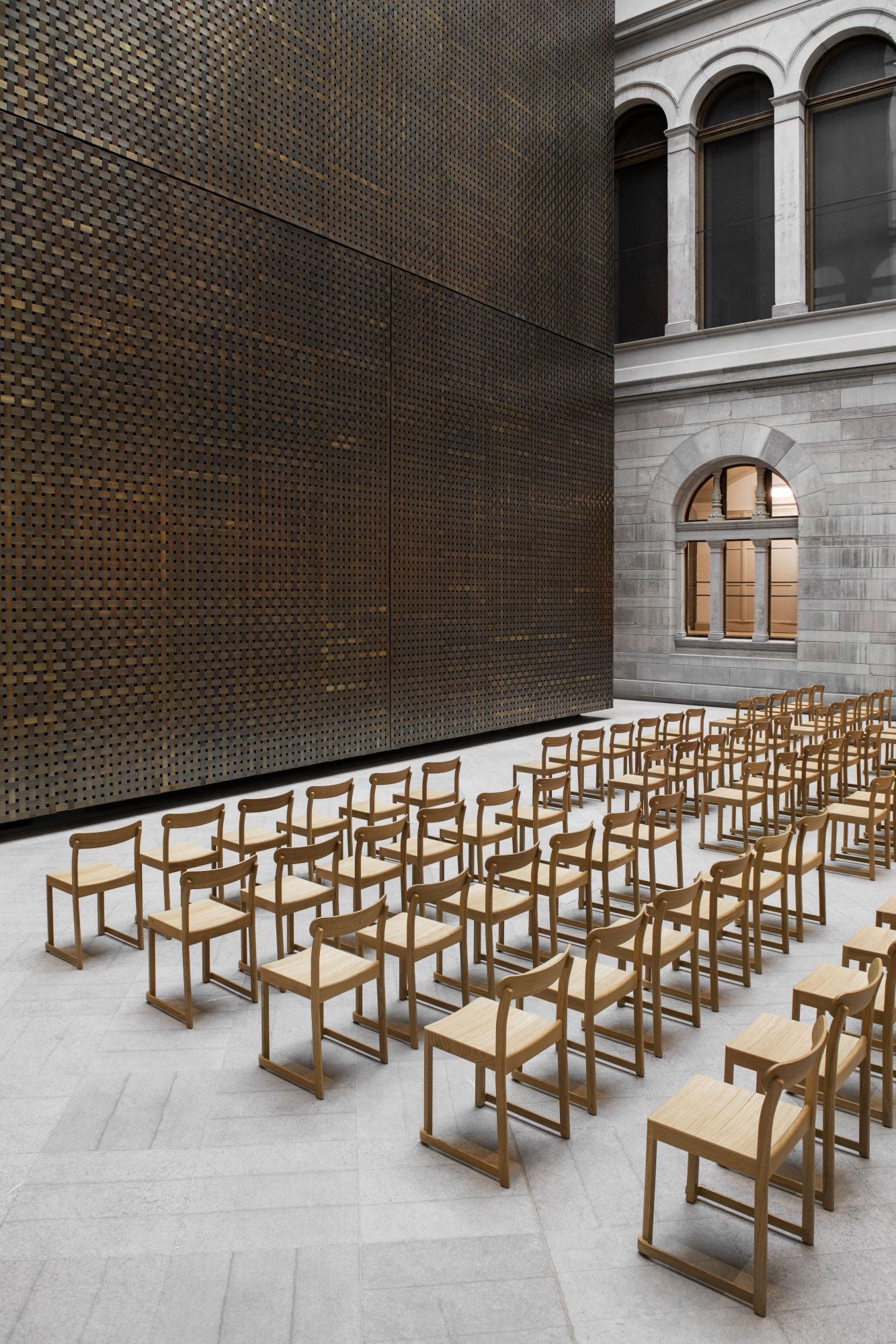 Row of wooden chairs in a courtyard with a modern metal wall and classical building facade.