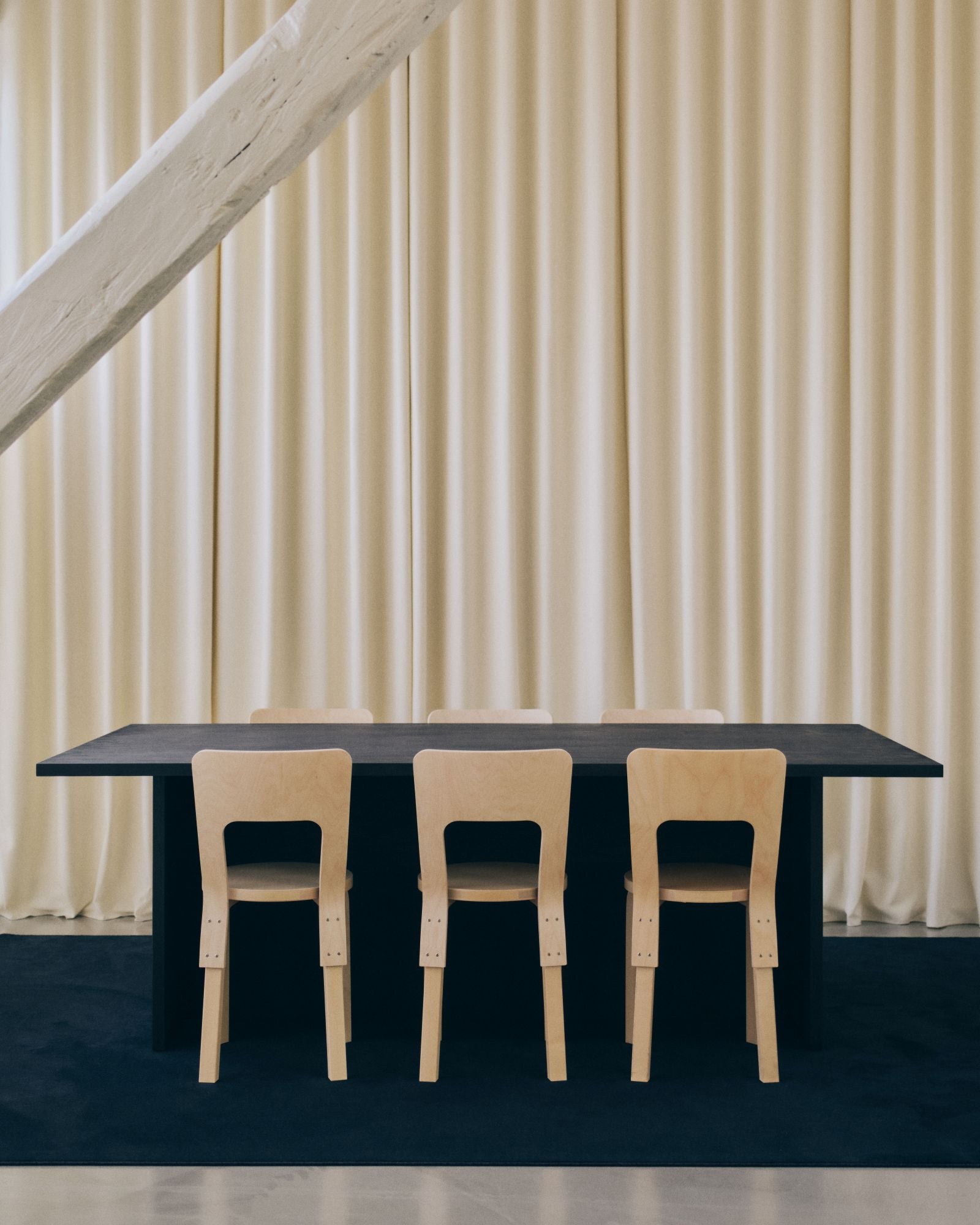 Long black table with four wooden chairs against a beige curtain background