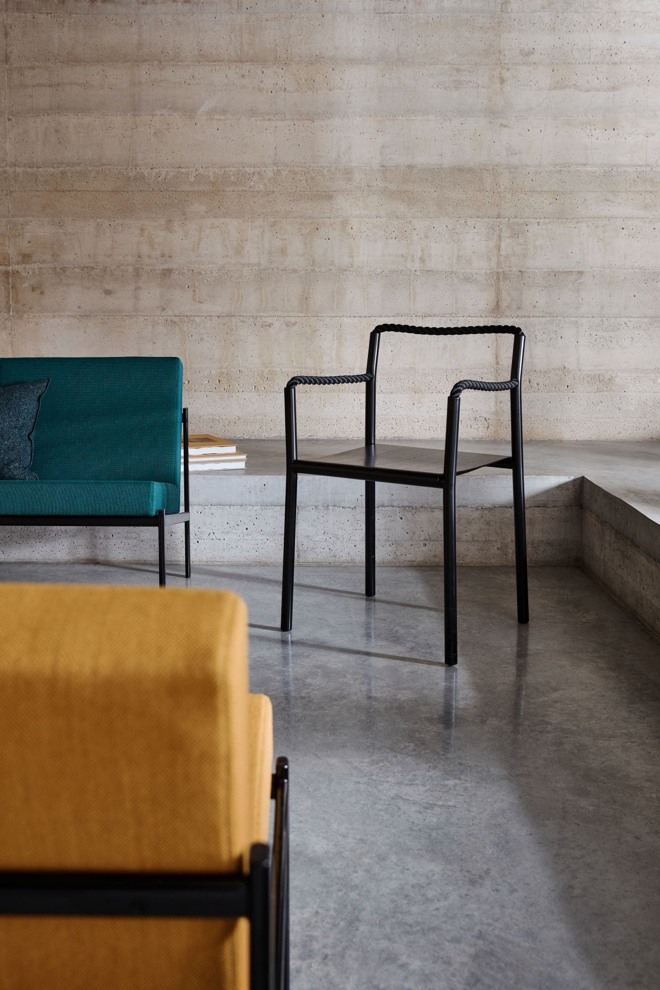 Modern furniture arrangement with a teal sofa, black chair, and yellow bench against a concrete wall.