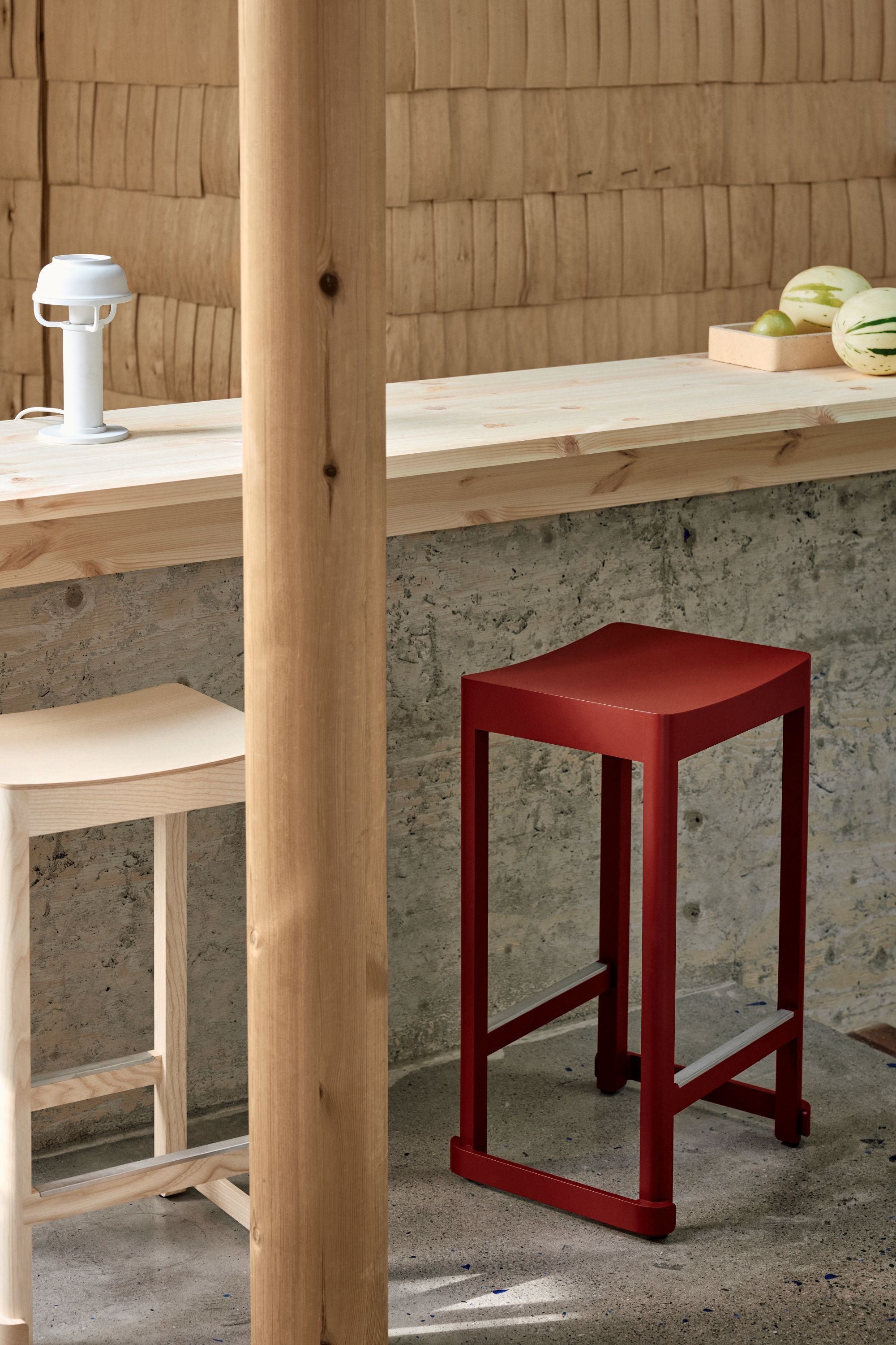 Red bar stool next to a wooden table with a concrete floor and wooden wall in the background.