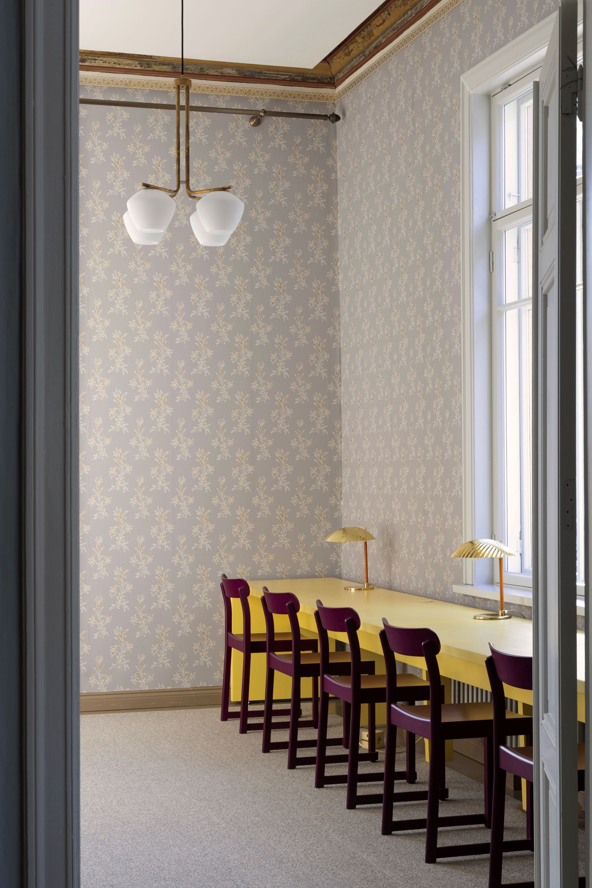 Long dining table with chairs in a room with patterned wallpaper and large windows.
