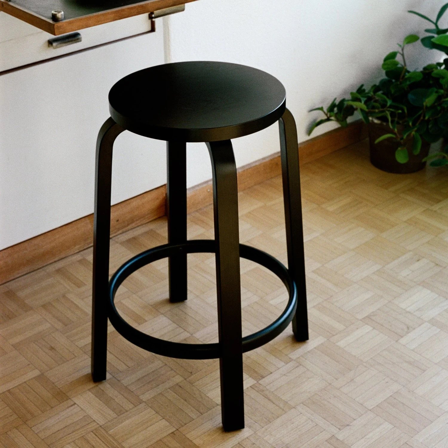 Black bar stool on a wooden floor with a white wall and plant in the background