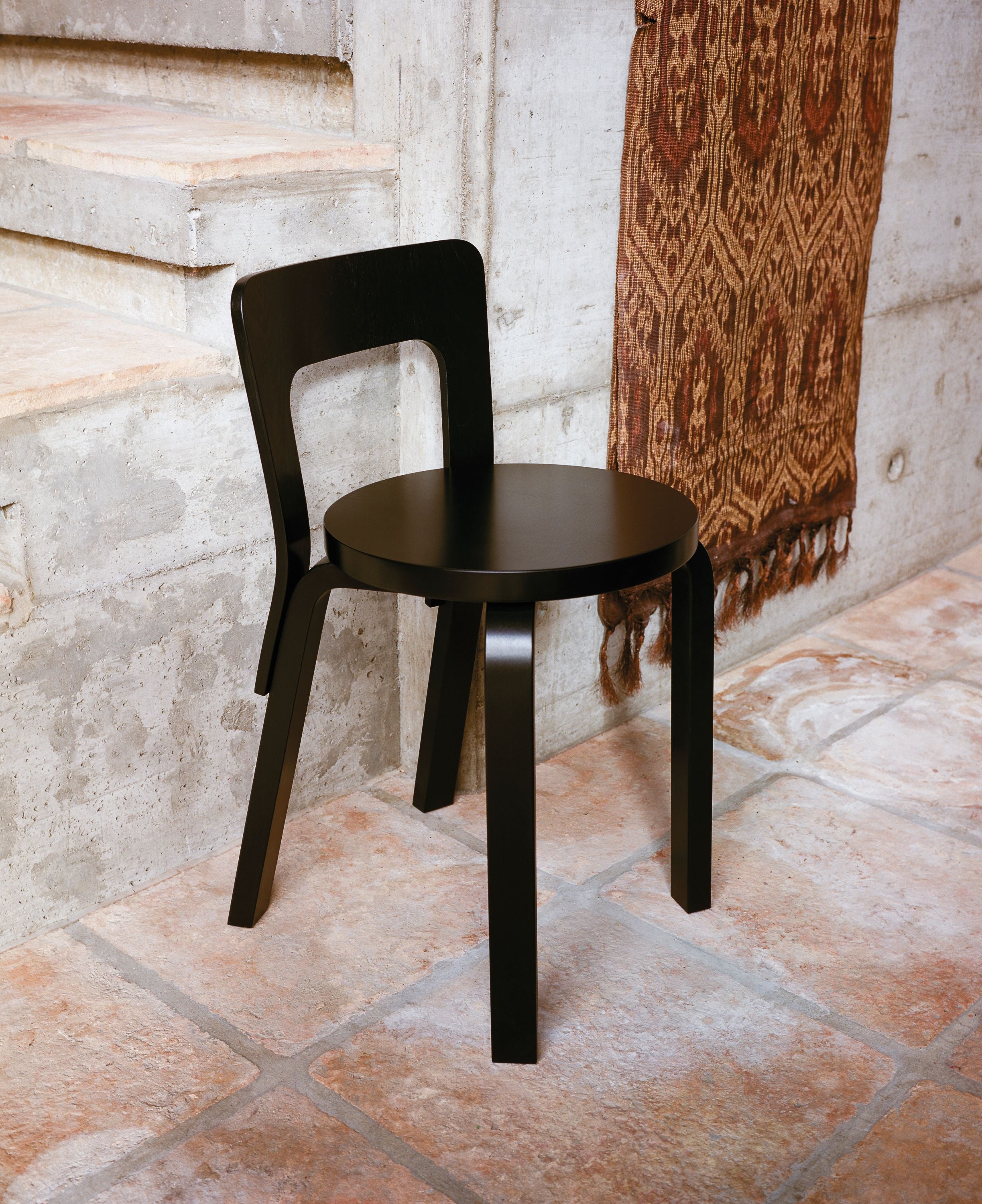 Black wooden chair on a stone floor with a textured wall and patterned fabric in the background.