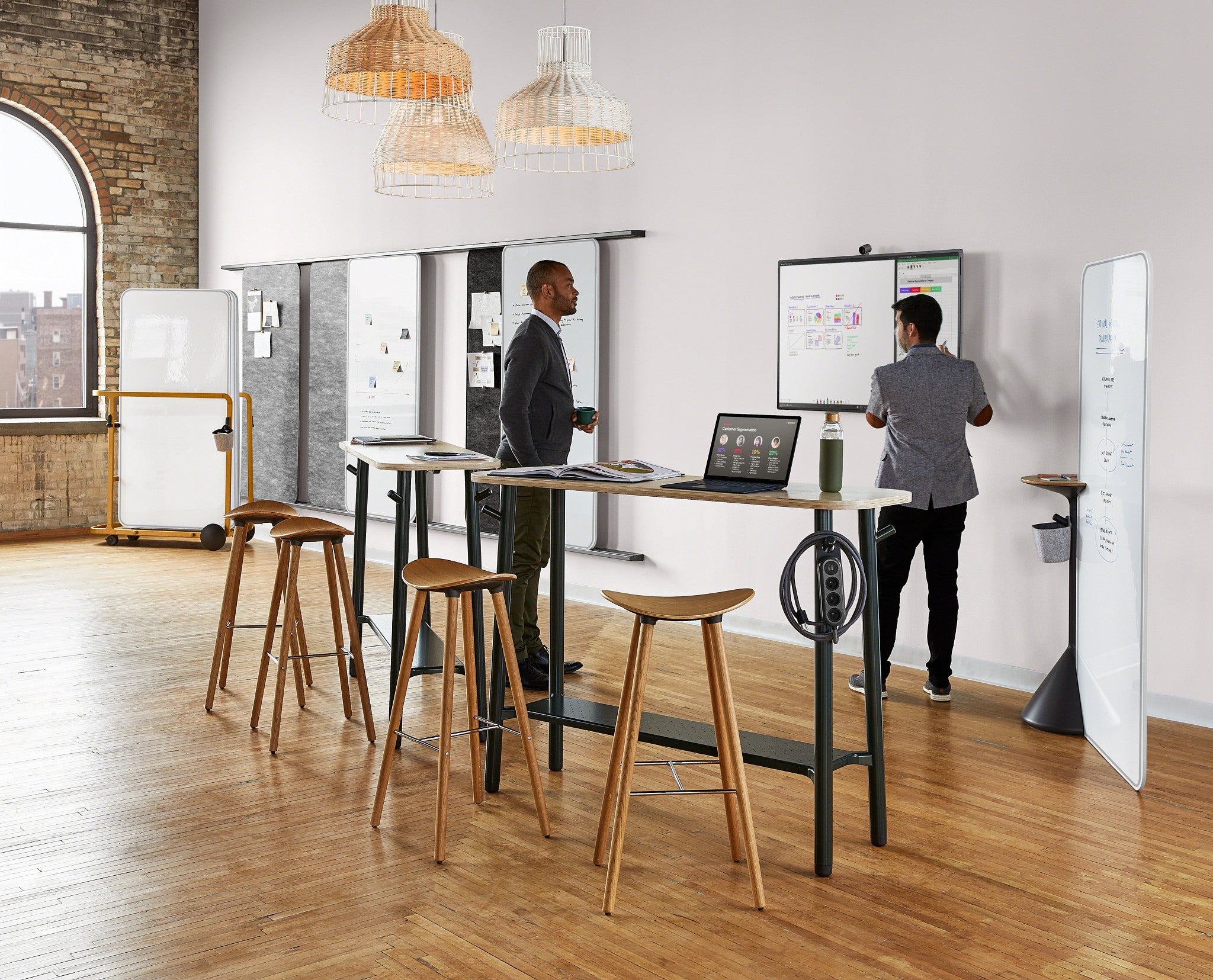 Two men working at a standing desk in a modern office setting with wooden floors and white walls.