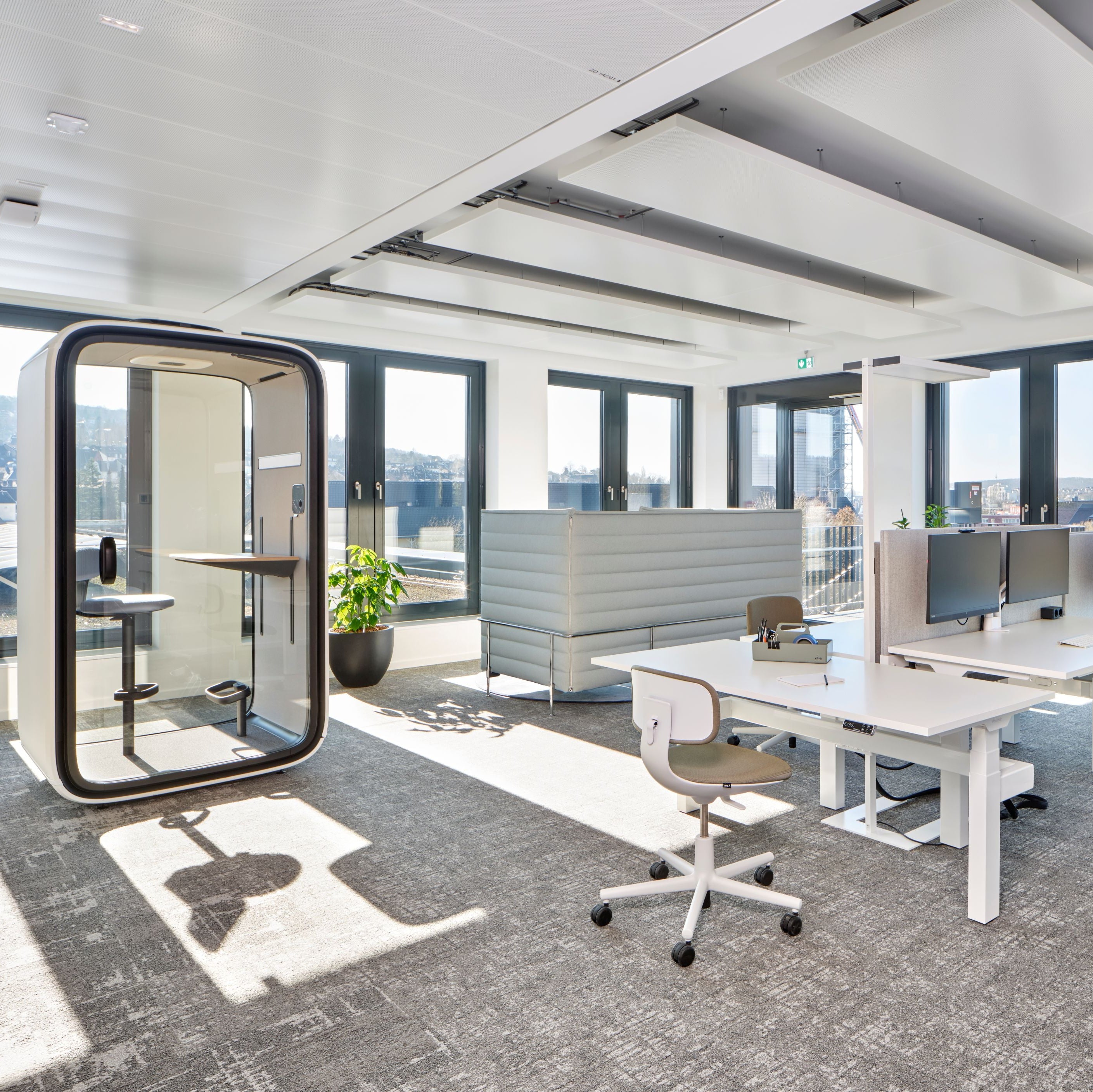 A modern open-plan office interior with white desks, swivel office chairs, large floor-to-ceiling windows, and a glass privacy call room on a gray carpeted floor.