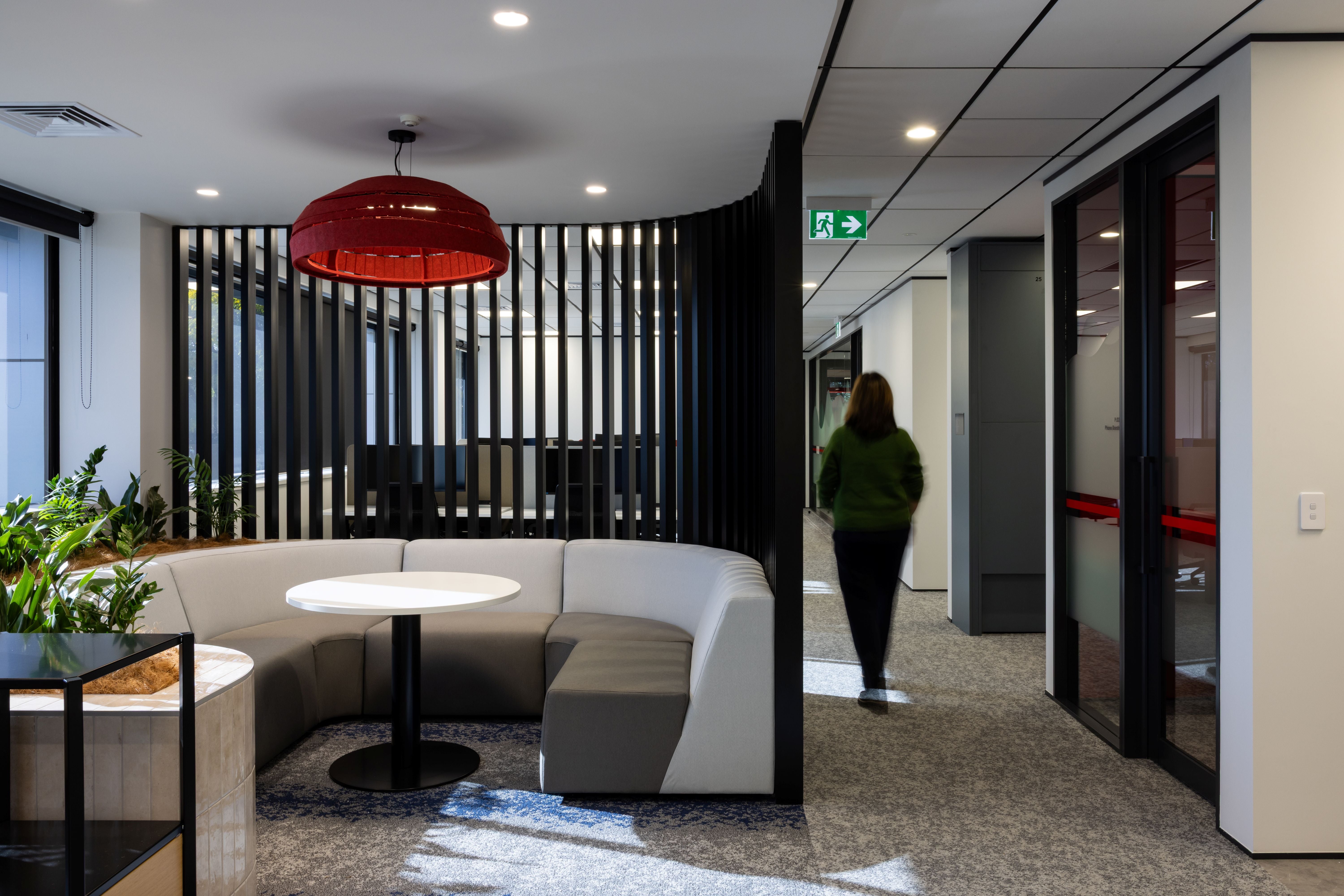 Modern office lounge area with a red pendant light, round table, and a person walking in the background.