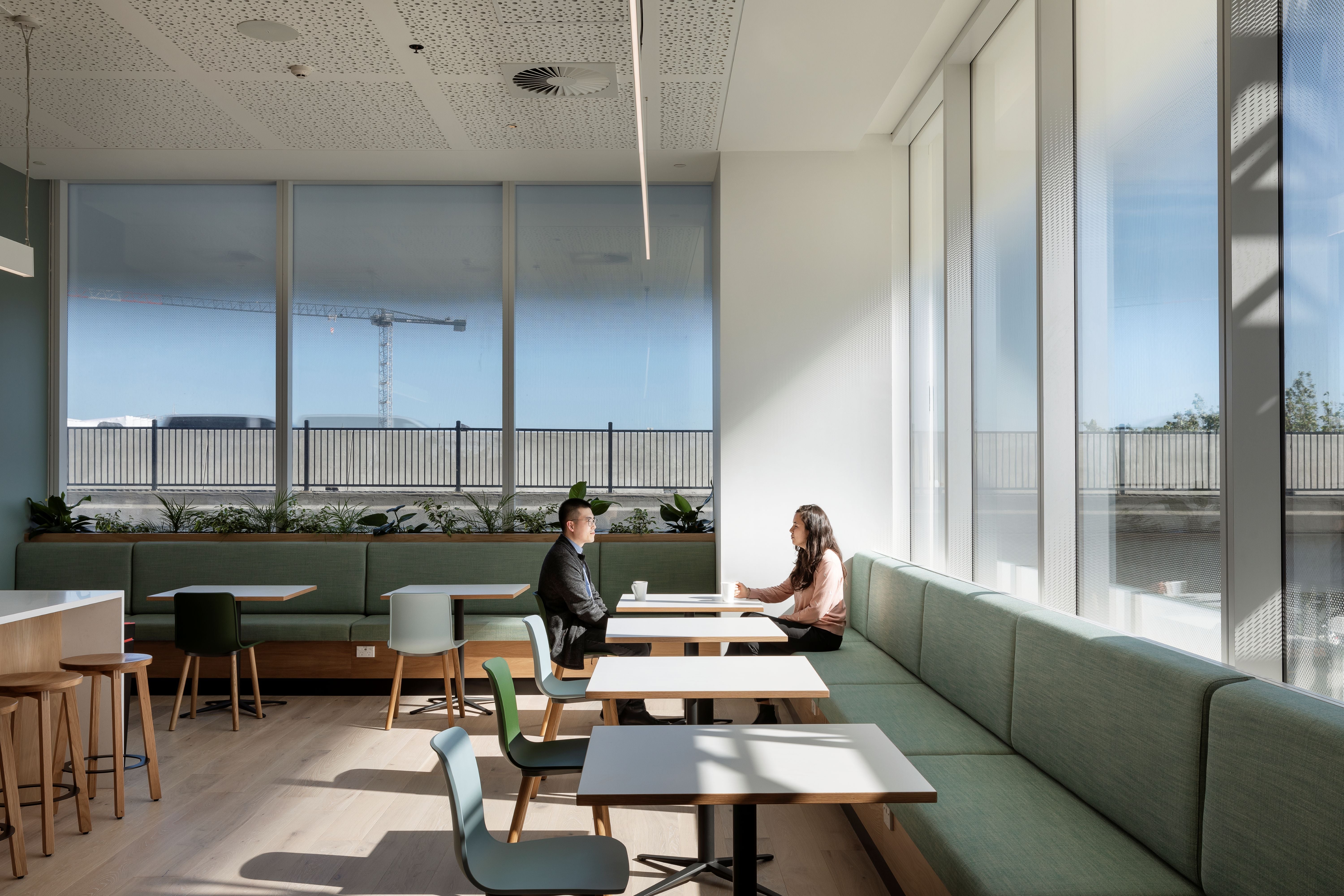 Modern office break room with large windows, people sitting at tables, and a view of a cityscape.