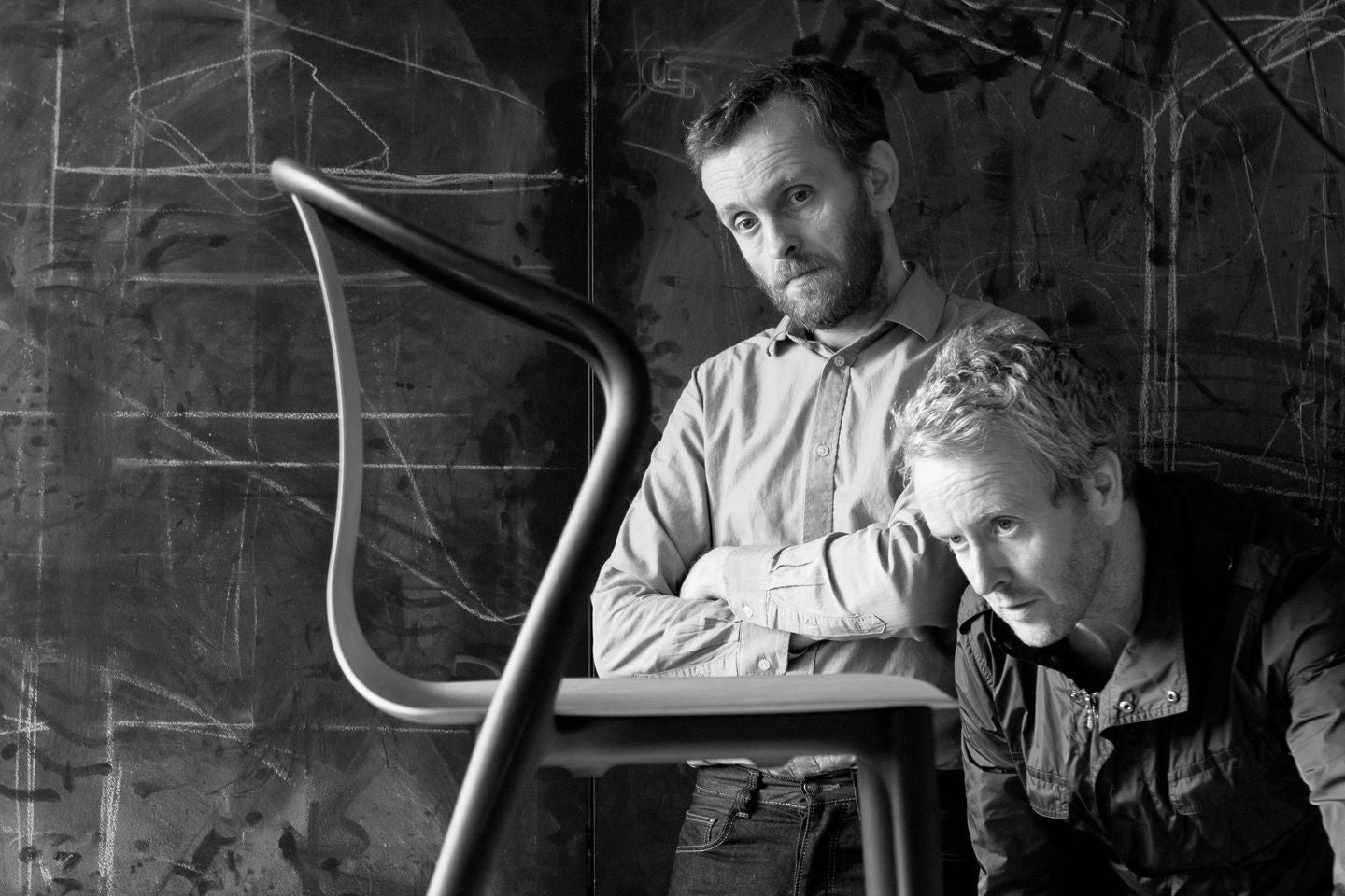 Two men sitting on a chair against a textured wall.