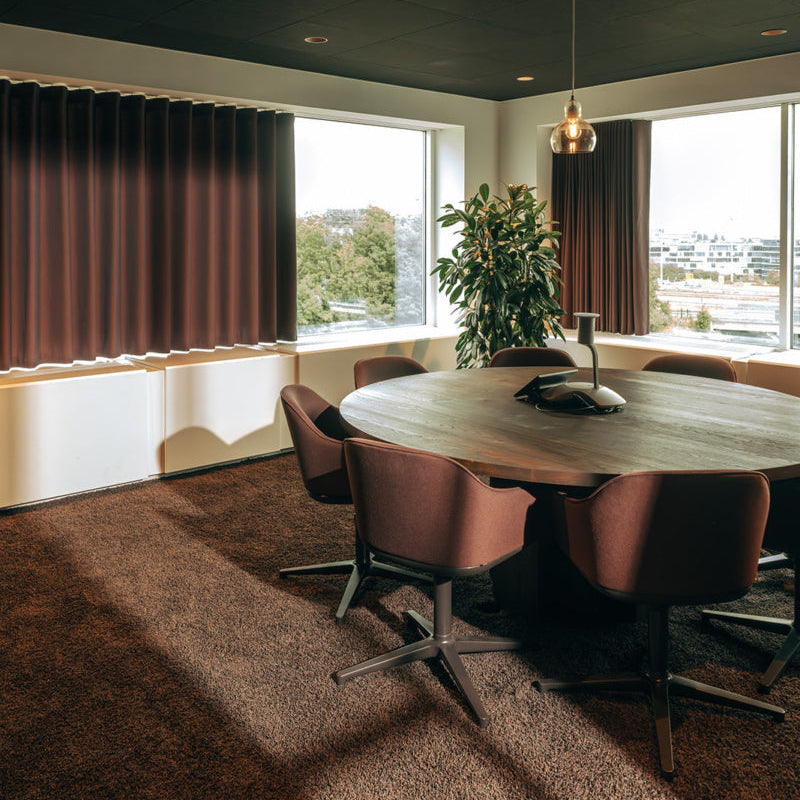 A modern meeting room featuring a round wooden dining table surrounded by brown upholstered chairs, set against large windows with dark brown pleated curtains and a dark ceiling.