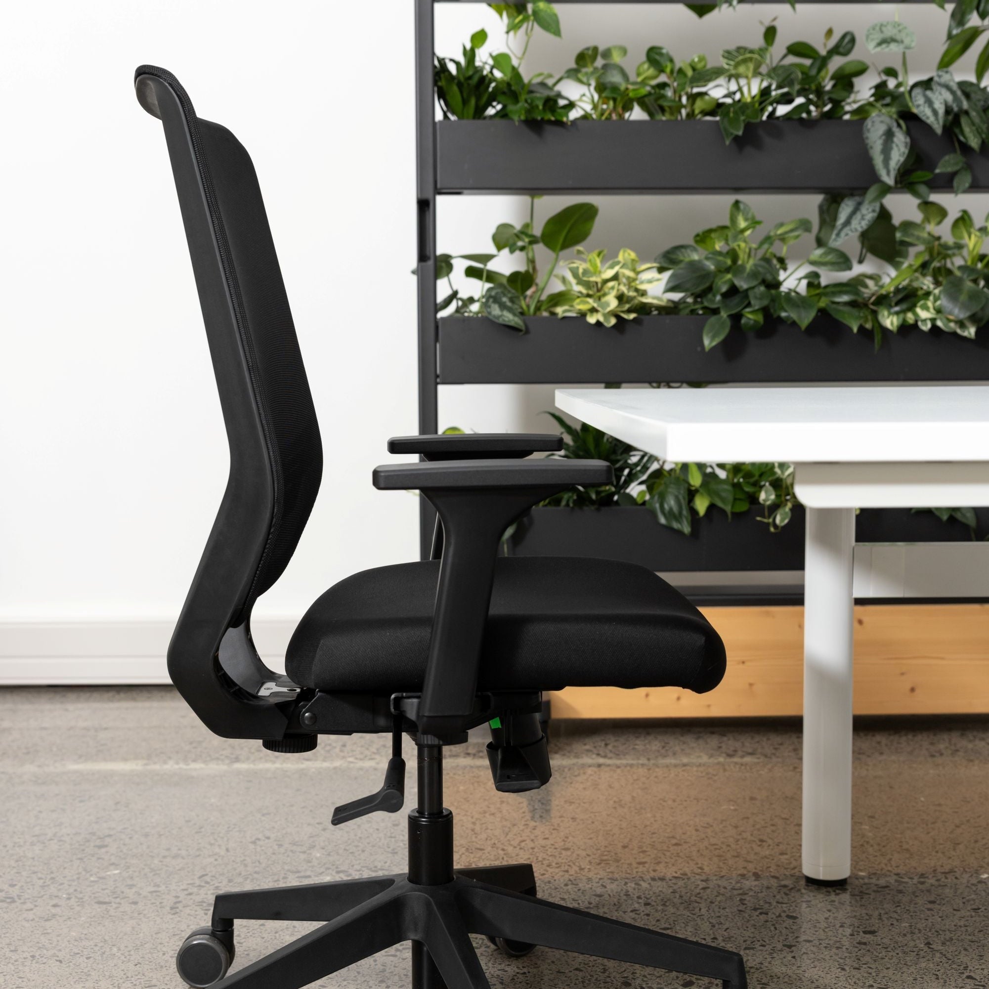 A modern black mesh office chair with a high back and five-wheel base, positioned next to a white desk in an office setting with a green vertical planter in the background.