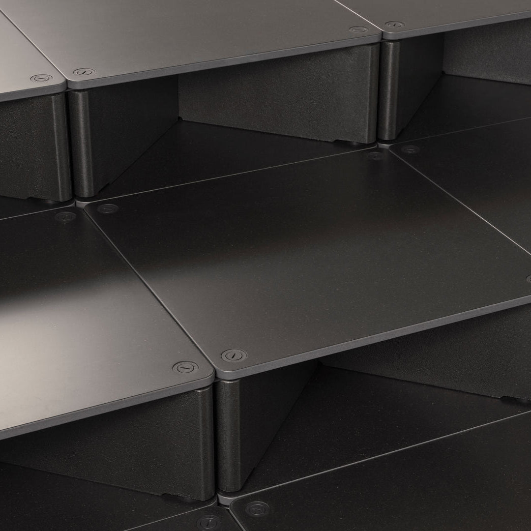 Close-up of dark gray modular storage boxes or shelves arranged in a geometric stack, showing matte panels and visible screw fasteners.