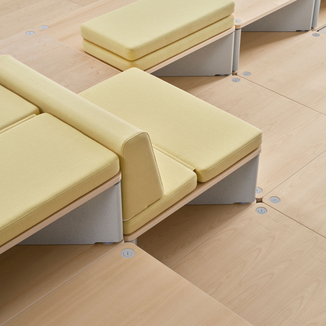 A close-up of modular upholstered seating with pale yellow seat cushions and light wood bases, arranged on a matching light wood floor in a clean interior setting.