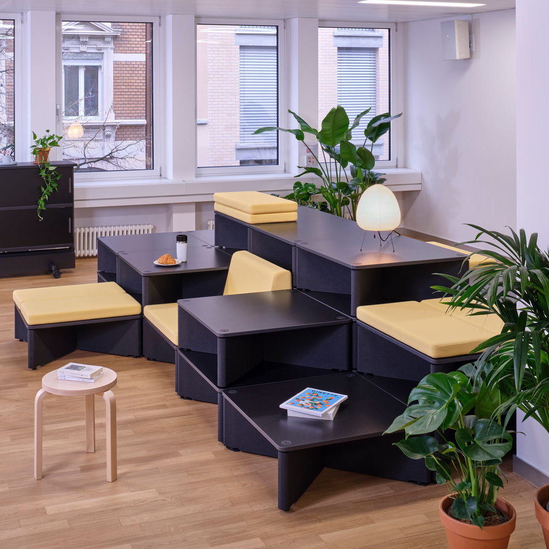 A modern modular lounge seating system with black geometric tables and benches featuring mustard yellow cushions, arranged in a bright office or lounge room with wood floors, large windows, and potted plants.