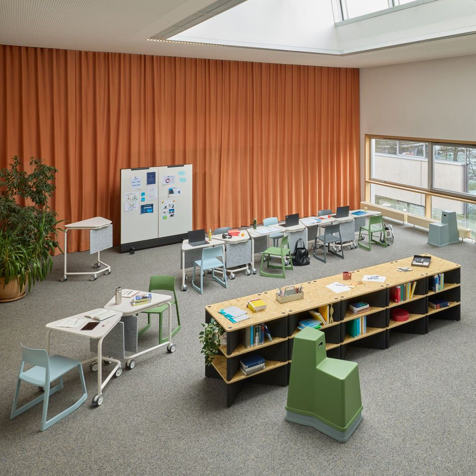 A bright modern classroom interior with multiple small desks, chairs, a long wooden bookshelf, whiteboards, and orange vertical wall panels. No people or animals are present, and the room appears to be shown as a product/interior design scene rather than a clean isolated product shot.